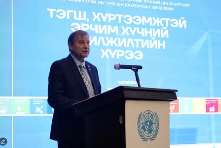 Man in suit speaks at podium bearing United Nations emblem; blue backdrop.