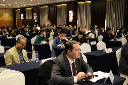 Photo: conference room filled with businesspeople at tables; foreground man in suit taking notes.