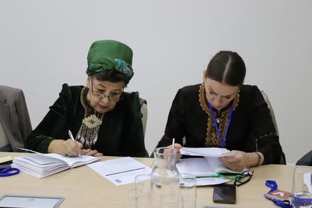 Three people sit at a table with papers and notebooks; one wears a green headscarf.