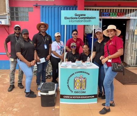 Group of people posing at a Guyana Elections Commission booth outdoors.