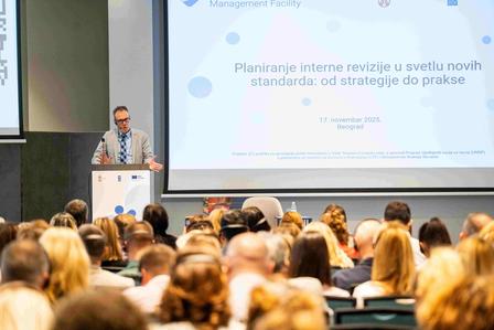 Photo of a speaker at a podium presenting to an audience in a conference hall with a slide.