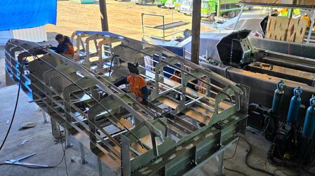 Long metal rack on a workbench in a workshop under a blue canopy; orange parts in slots.