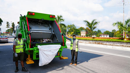 Two sanitation workers in reflective vests load a white bag into a green garbage truck on a road.