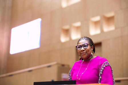 Speaker in pink blouse at podium in a wood-paneled conference hall.