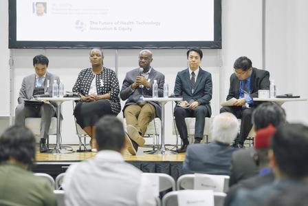 Six panelists seated on stage at a conference, audience in foreground.