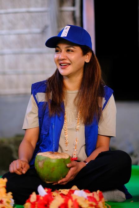Person wearing blue cap and blue vest sits cross-legged on a green mat, holding a green coconut.
