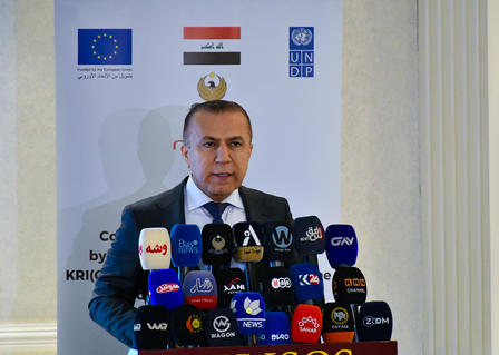 Speaker at a press conference podium with numerous microphones; EU and Iraqi flags in the background.
