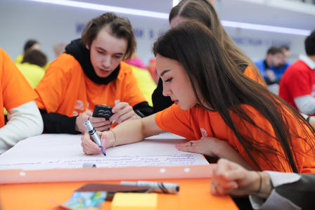Students in orange shirts sit at a table; a girl writes on a large sheet of paper with markers.