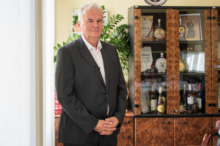 Man in a dark suit stands with hands folded beside a glass-fronted china cabinet in a bright room.