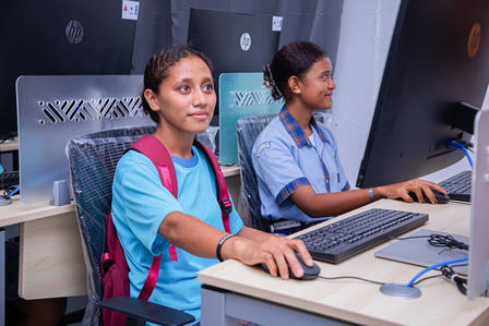 Student with backpack at a desktop computer beside a teacher in a classroom computer lab.