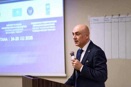 Man in a suit speaks at a podium with a microphone in a conference room; projector screen behind.