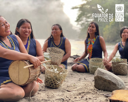Women sit in a circle on a rocky beach; one plays a drum as woven baskets sit nearby, with a logo in the corner.