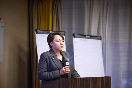 Speaker at a wooden podium giving a presentation, with flip charts in a conference room.