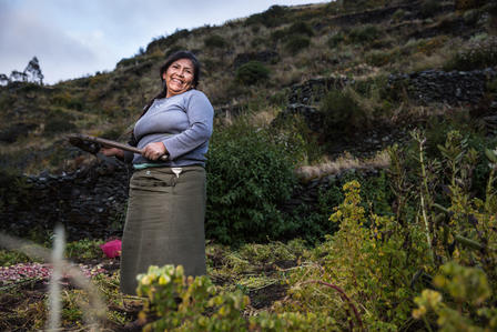 Smiling woman in a garden holding a long-handled tool, with flowers and hills behind.
