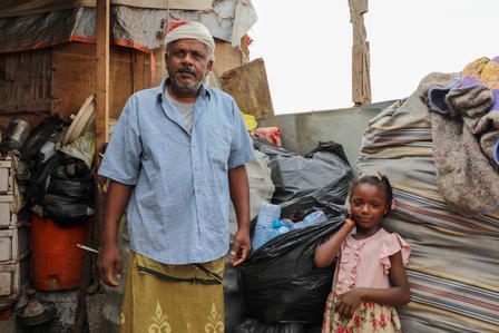Photograph of a man in a light-blue shirt with a young girl amid makeshift shelters.