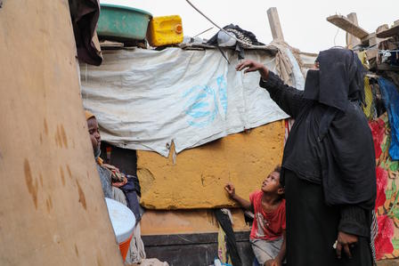 Displaced family at a damaged shelter with blue-tarp walls; woman in black hijab beside a child.