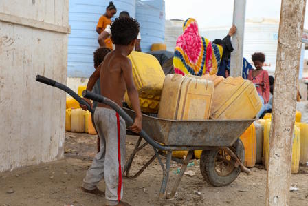 Person pushes a wheelbarrow loaded with yellow jerrycans among people in a dusty outdoor setting.