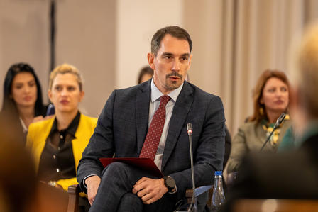 Panelist in a dark suit and red tie sits on stage, holding a red folder.
