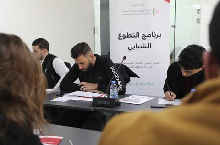 Participants at a workshop, writing on sheets, with an Arabic banner in the background.