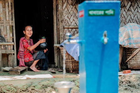 Two women sit on a wooden village porch beside a tall blue water dispenser.