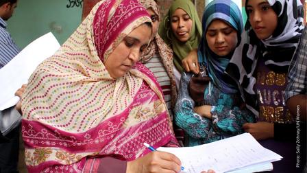 Woman in pink patterned dress and headscarf writes on a sheet at a table, with others nearby.