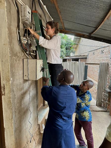 Three people fix outdoor electrical wiring on a brick wall beneath a corrugated metal roof.