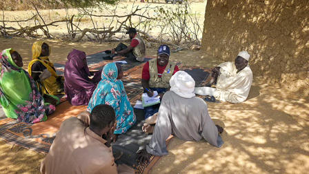 Group of women in colorful clothing sitting in a circle under a tree in a dusty outdoor setting.