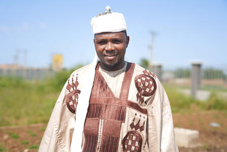 Person wearing a cream and brown patterned robe stands in an open field under a clear blue sky.