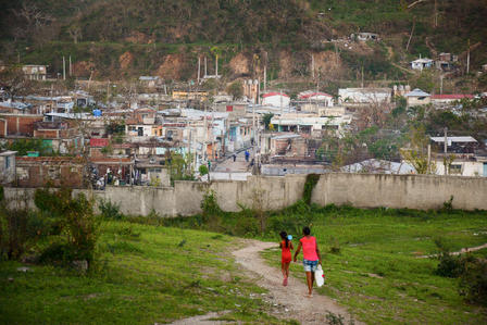 Two people walk a dog along a grassy path toward a hillside town of many small houses.