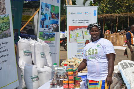 Woman in white T-shirt and colorful wrap at an outdoor exhibit booth with posters and stacked bags.
