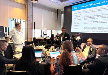 Photograph of a conference room meeting, attendees around a table while a presenter shows slides.