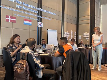 Photograph of a conference room with several people around a table; Denmark, Netherlands flags.