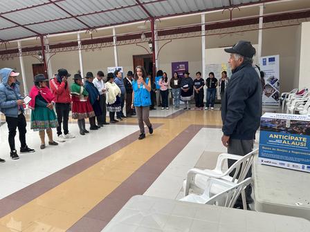Photograph of a man in a hat addressing a line of people in a hall with tables and banners.