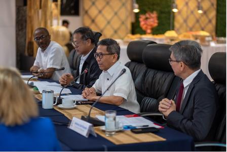 Photograph of a formal business meeting with several professionals around a conference table, microphones visible.