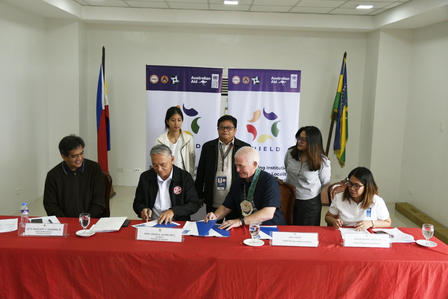 Photograph of officials signing documents at a long red table, flags in the background.