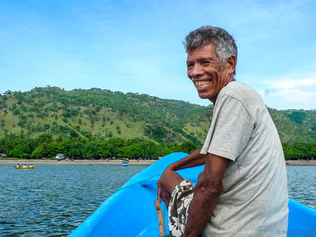Man standing on a blue boat on calm water with green hills in the background.