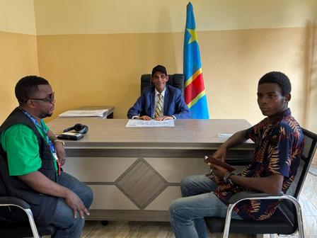 Three men in an office; official at desk with blue-yellow-green flag behind, two visitors seated.
