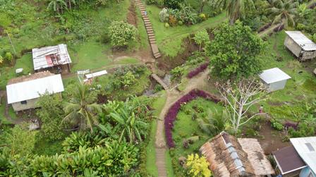 Aerial view of a tropical village with palm trees, dirt roads, purple path, and tin-roof houses.