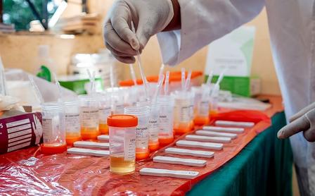 Lab scene with a gloved hand using a pipette over orange-capped vials on a red table.