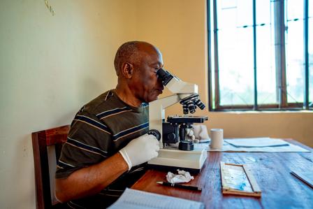 Man with striped shirt and bandaged arm sits at a table in a bright room beside a microscope.