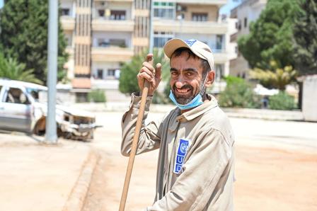 Man in beige tunic holding a wooden staff on a dusty street, apartment buildings in the background.