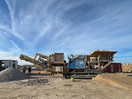 Construction site with large orange machinery, a conveyor, and gravel piles under a blue sky.