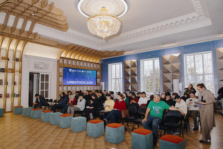 Photo: Conference in a grand hall with chandeliers; audience on blue ottomans listening.