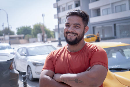 Man with crossed arms in an orange shirt on a street with cars and buildings behind.