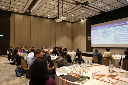 Audience at round tables in a conference room watches a presenter with a projected slide.