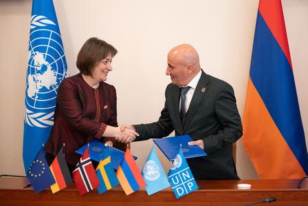 Photograph of two officials shaking hands across a table with flags, including a UN flag.
