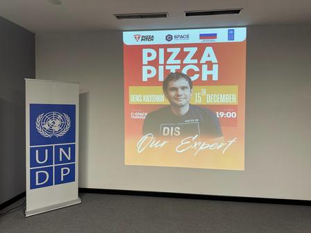 Conference room with an UNDP banner; projection displays a Pizza Night poster featuring a man.