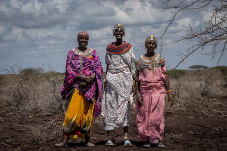 Three African women in vibrant traditional dresses stand on a dusty plain beneath a cloudy sky.