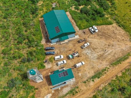 Aerial view of teal-roofed buildings with vehicles on a dirt lot amid green surroundings.