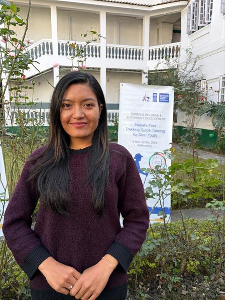 Person in maroon sweater stands outdoors near a sign in front of a white building.